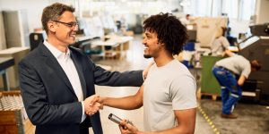 Happy manager shaking hands with African American factory worker in industrial building.