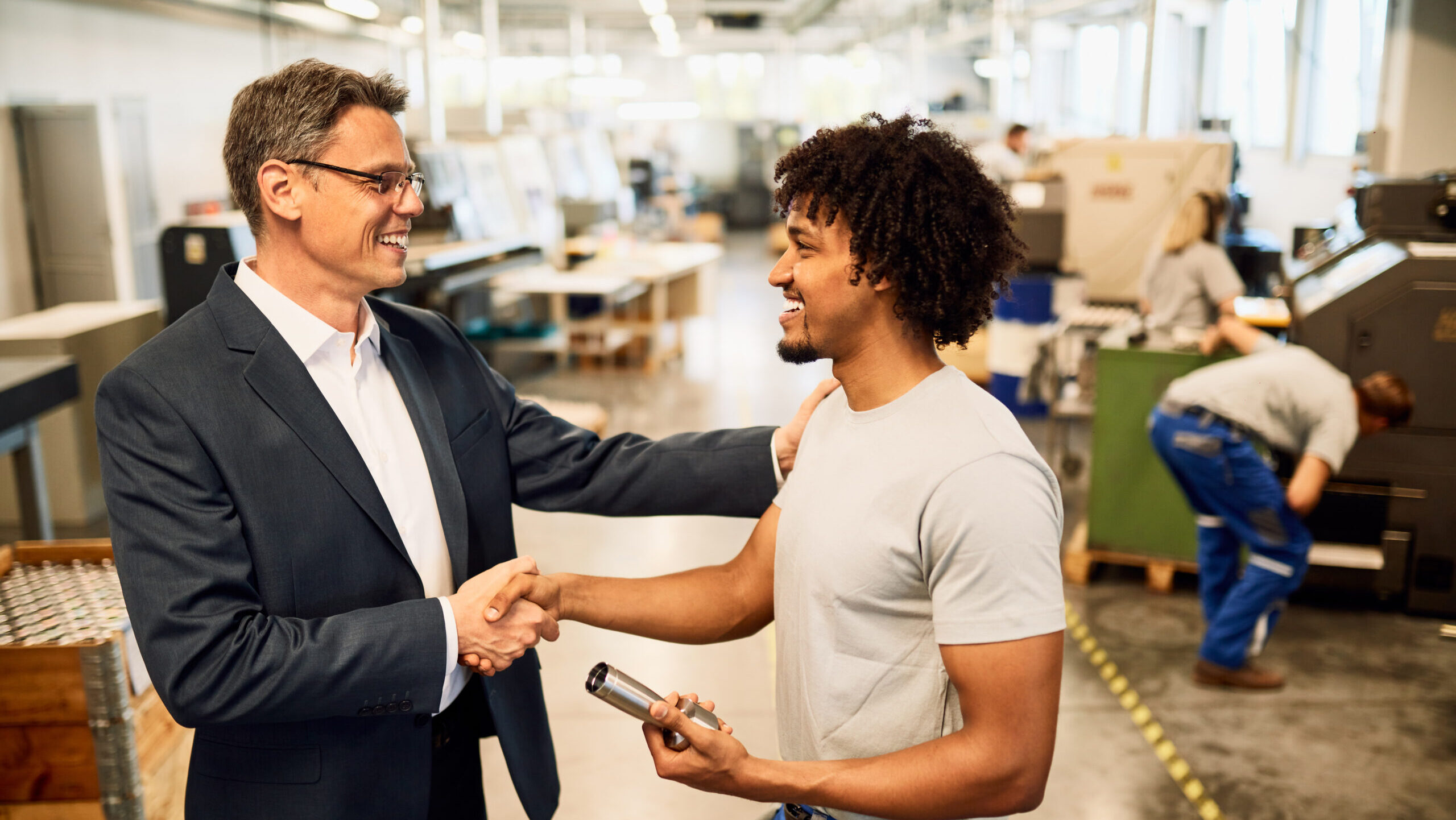 Happy manager shaking hands with African American factory worker in industrial building.